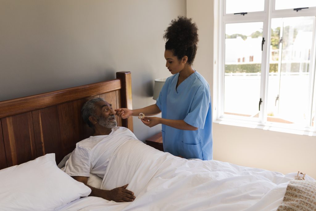 Front view of a young African American female doctor fitting a senior African American man with hearing aid in bedroom at home